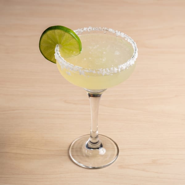 A margarita in a coupe glass with a salted rim and a lime wedge on a light wooden surface.