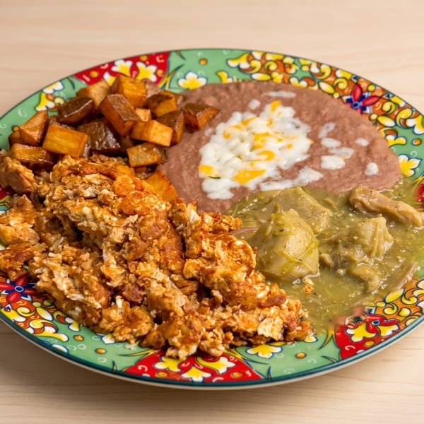 A colorful plate of scrambled eggs, refried beans with cheese, green chili, and roasted potatoes.