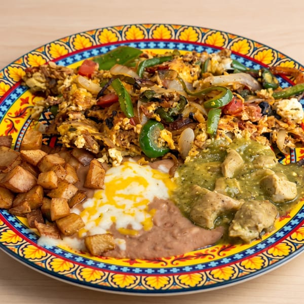 A colorful plate of scrambled eggs with peppers and onions, potatoes, beans, and green chili chicken.