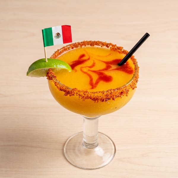 Mango margarita with a chili-lime rim, lime wedge, and a small Mexican flag on a light wooden table.