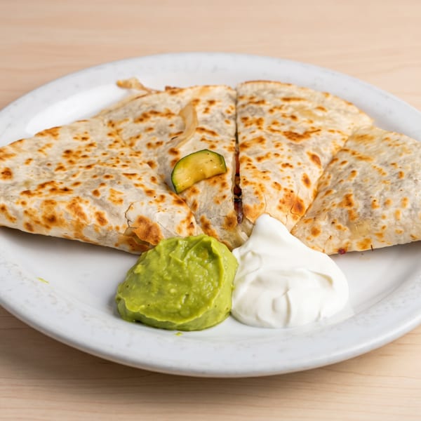 A quesadilla cut into four triangles, served with guacamole, sour cream, and a zucchini slice.